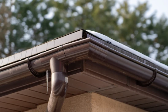 Détail photographique en gros plan d'une toiture pendant des travaux d'étanchéité, montrant l'application ou la pose d'une membrane d'étanchéité (bitumineuse ou synthétique) sur la pente d'une toiture. Matériau gris, noir ou blanc clairement visible, fixation mécanique ou collage en cours. Surface de la toiture légèrement mouillée ou humide, suggérant les conditions d'un environnement extérieur. Mains d'un ouvrier qualifié en gants de travail manipulant ou lissant le matériau. Environnement de jour naturel, éclairage rasant soulignant les détails de texture. Cadrage professionnel et technique mettant l'accent sur la qualité et la rigueur du travail d'étanchéification, sans texte ni logo visible.